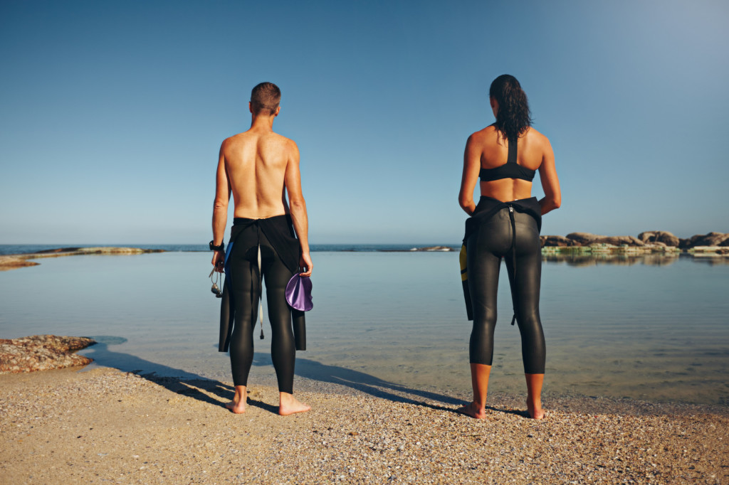 Rear view of young athletes standing on lake preparing for triathlon. Man and woman preparing for a race wearing a wetsuit.